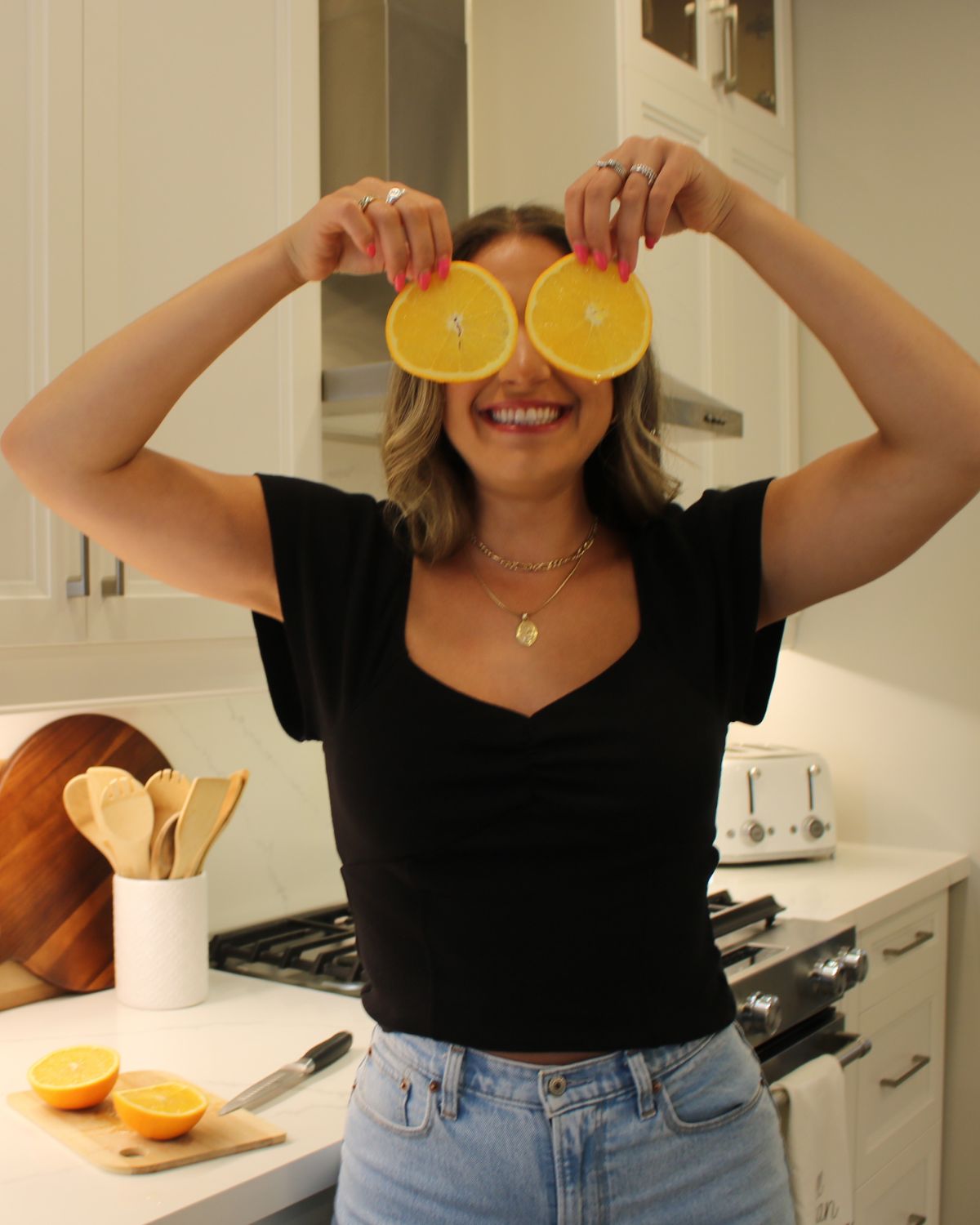 registered dietitian standing in kitchen - wearing jeans and black t shirt holding up to orange slices on over her eyes 