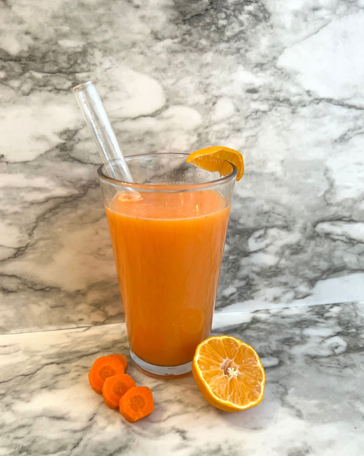 glass cup filled with Orange Ginger Carrot Juice  with glass straw instead - slice of orange on the cup sliced carrots in front front of cup with half an orange. 