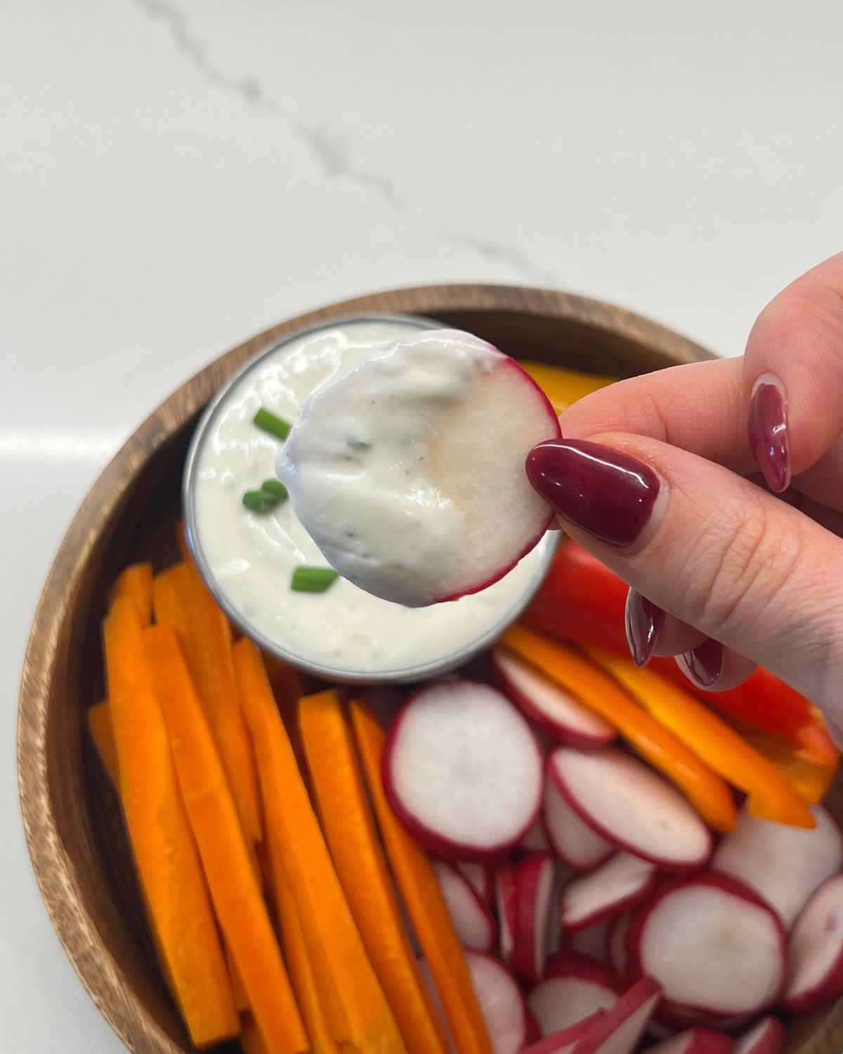 ranch dip in a bowl inside a bigger bowl with chopped carrots, peppers and radish - holding up radish dipped in ranch 